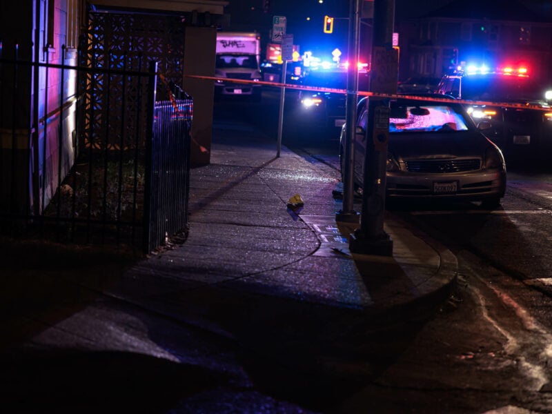 A nighttime scene on a suburban street. Police vehicles display red and blue flashinging lights in the background. The foreground shows crime scene tape and, behind that, a car with a shattered window.