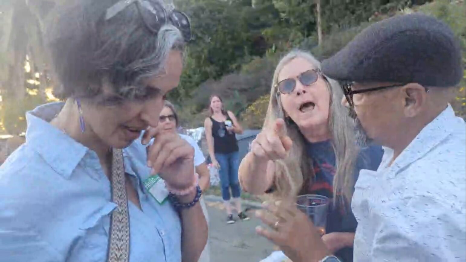 Group of three people engaged in an animated discussion outdoors during an event. The person in the center gestures emphatically towards the camera. They are surrounded by other attendees in a casual, sunlit setting.