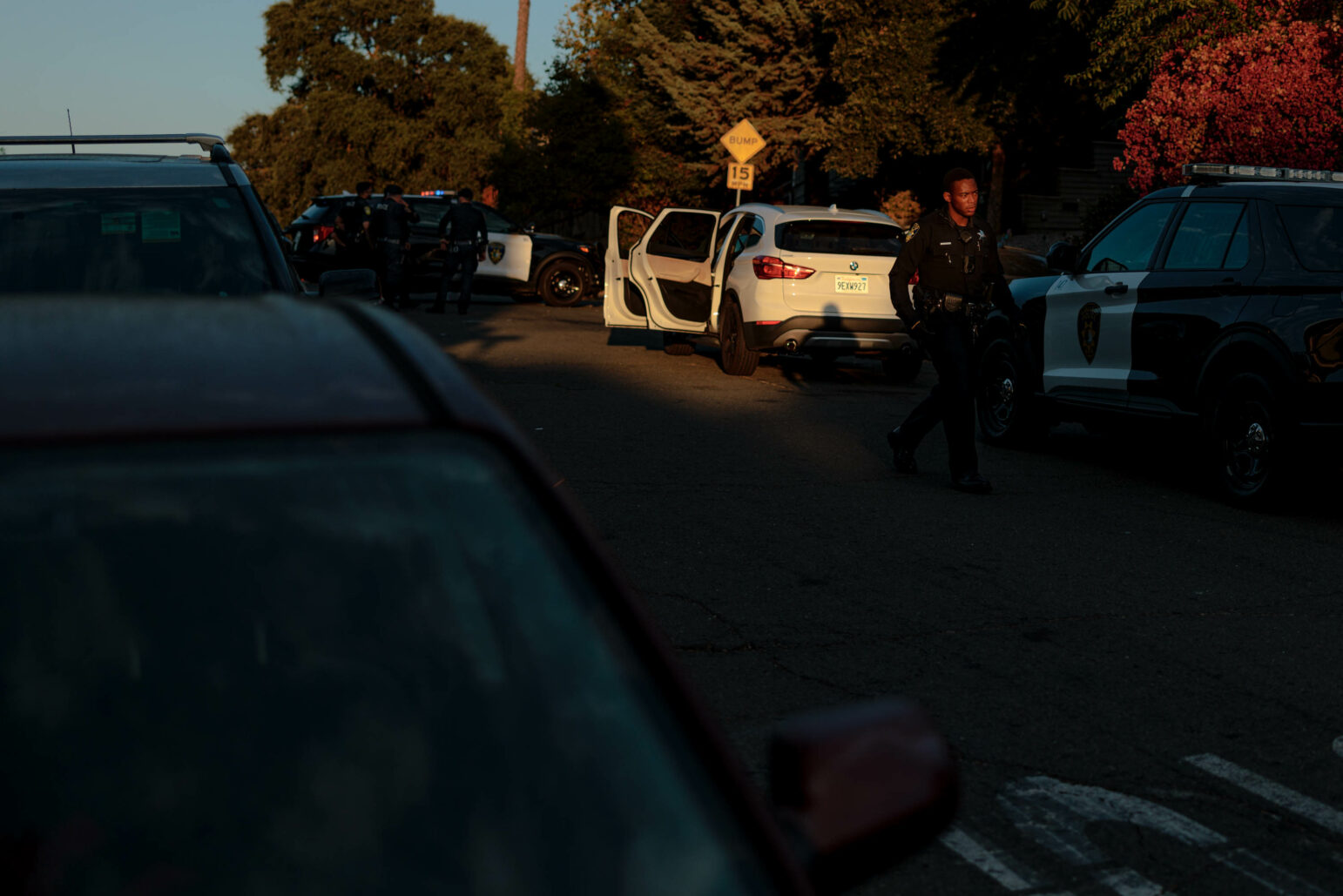 A street scene at dusk showing multiple police vehicles, including a black-and-white patrol car. Several police officers are gathered near the vehicles, with one officer walking towards the foreground. The area is lit by the setting sun, casting long shadows. A white SUV is parked in the middle of the scene, with its driver's door open. The atmosphere suggests a police investigation or search operation.