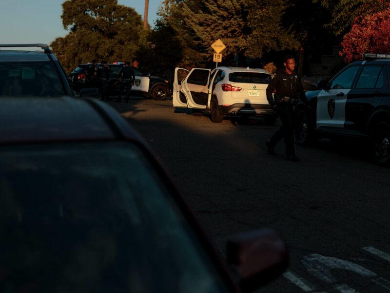 A street scene at dusk showing multiple police vehicles, including a black-and-white patrol car. Several police officers are gathered near the vehicles, with one officer walking towards the foreground. The area is lit by the setting sun, casting long shadows. A white SUV is parked in the middle of the scene, with its driver's door open. The atmosphere suggests a police investigation or search operation.