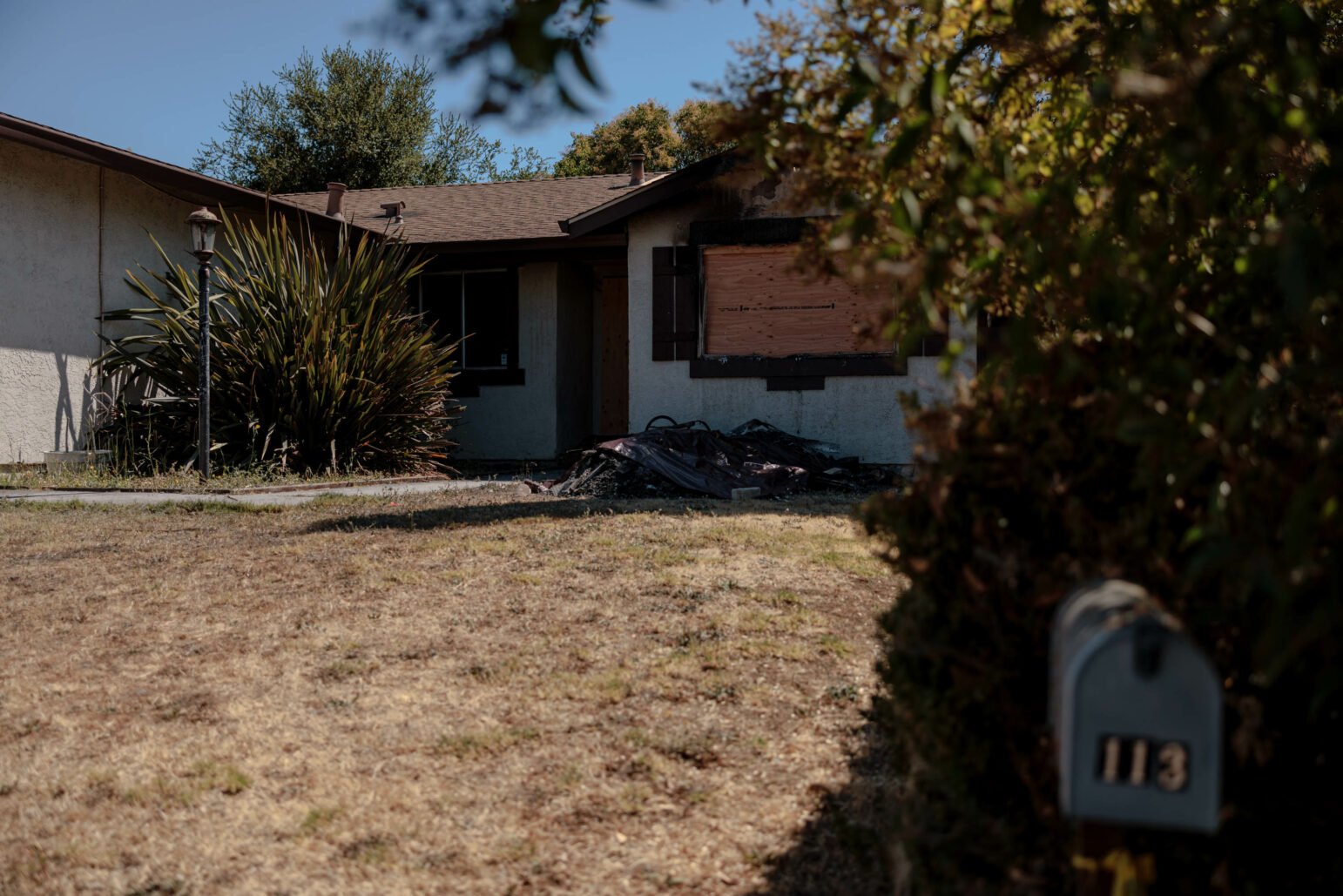 A single-story suburban house with beige stucco walls and a brown roof. The front yard is dry, with a patchy lawn and a few shrubs, including a tall plant near the front of the house. The front door and one of the windows are boarded up, and there is a pile of burnt debris in front of the house, suggesting damage from a recent fire. A mailbox with the number "113" is visible in the foreground, partially.