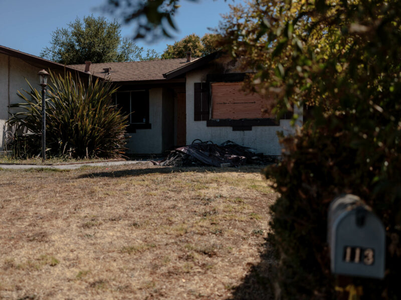 A single-story suburban house with beige stucco walls and a brown roof. The front yard is dry, with a patchy lawn and a few shrubs, including a tall plant near the front of the house. The front door and one of the windows are boarded up, and there is a pile of burnt debris in front of the house, suggesting damage from a recent fire. A mailbox with the number "113" is visible in the foreground, partially.