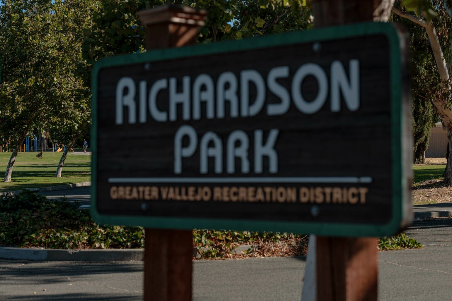 A close-up of a wooden sign for "Richardson Park" with white lettering and a green border, partially obscuring the background. The sign also includes smaller text that reads "Greater Vallejo Recreation District." In the background, there is a grassy area with trees and a person playing with a dog in the park. The overall scene suggests a quiet, sunny day in a well-maintained public park.