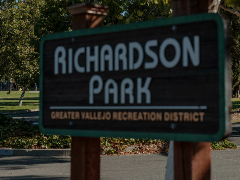 A close-up of a wooden sign for "Richardson Park" with white lettering and a green border, partially obscuring the background. The sign also includes smaller text that reads "Greater Vallejo Recreation District." In the background, there is a grassy area with trees and a person playing with a dog in the park. The overall scene suggests a quiet, sunny day in a well-maintained public park.