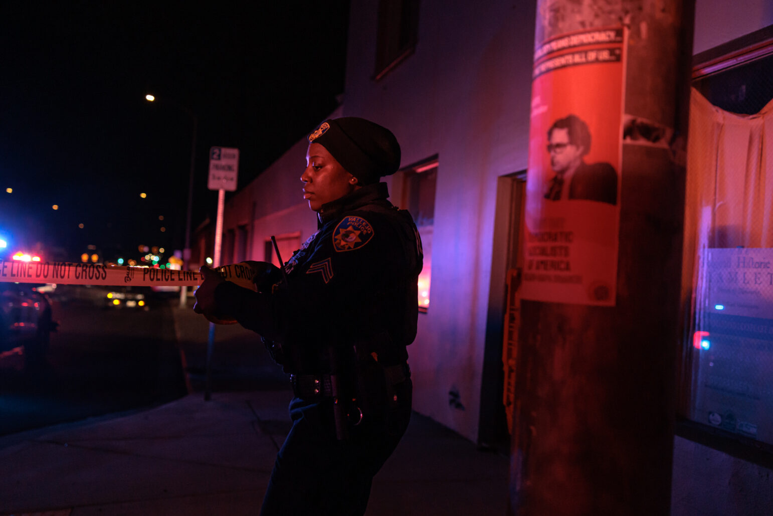 A female police officer, dressed in a dark uniform and beanie, carefully holds a "Police Line Do Not Cross" tape as she secures a crime scene at night. The scene is lit by the red and blue flashing lights of nearby police vehicles.