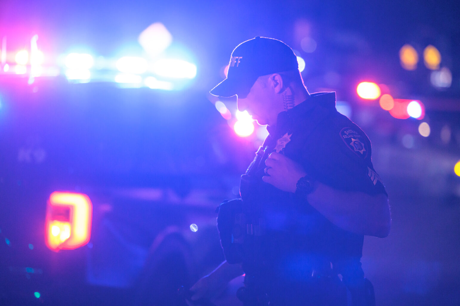 A sheriff's deputy, wearing a black uniform and hat, is illuminated by the blue and red lights of patrol vehicles at night, creating a dramatic contrast between the bright lights and the dark surroundings.