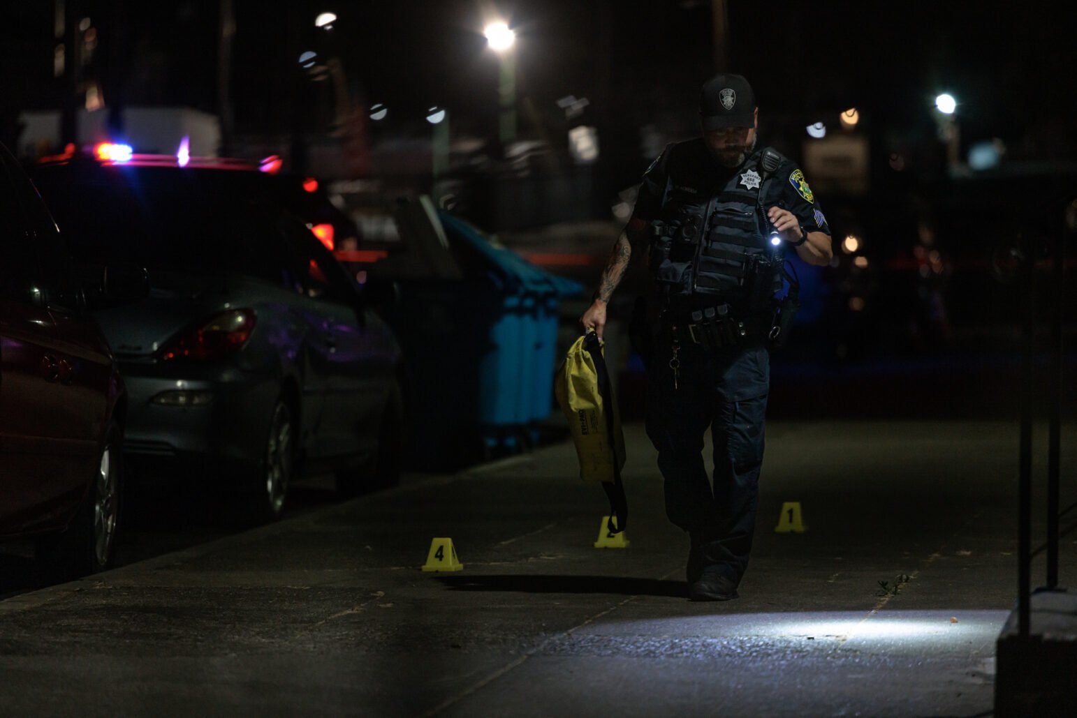 A police officer walks along a dimly lit sidewalk at night, marked with yellow evidence markers. The scene is illuminated by the officer's flashlight and the distant glow of police vehicle lights