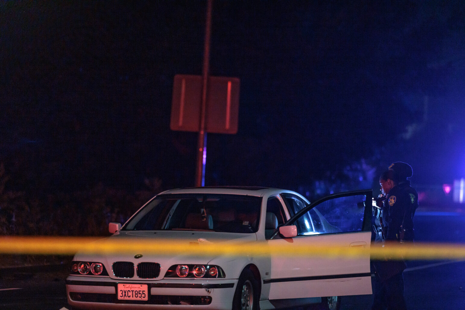 Two police officers stand next to a white BMW sedan at night, with the car door open. The scene is part of a larger crime scene, with yellow tape marking the area.