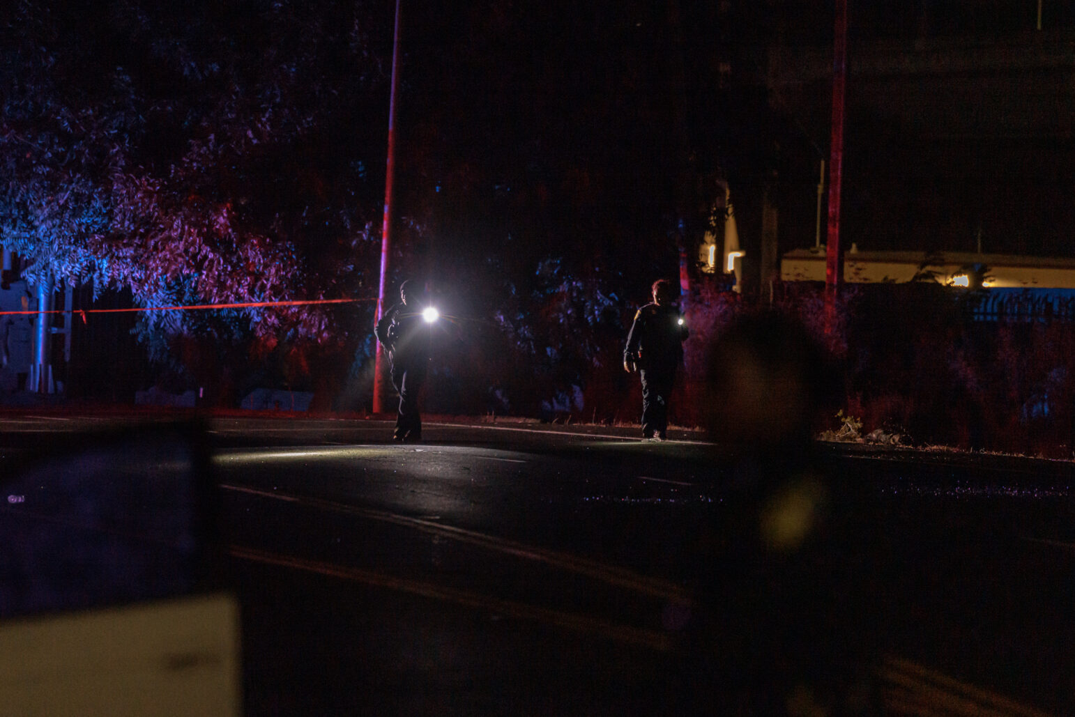 Two officers shine flashlights as they walk down a dark road, which is cordoned off with crime scene tape. The area is dimly lit, with the blue and red flashing lights in the distance casting shadows on the road.