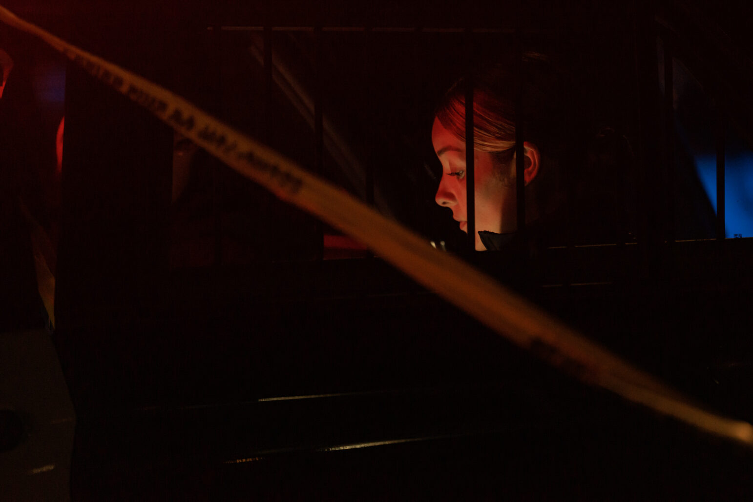 A police officer in a car at night is partially obscured by yellow crime scene tape, with only her face visible, lit by the red and blue lights outside the vehicle. The image captures a moment of concentration.