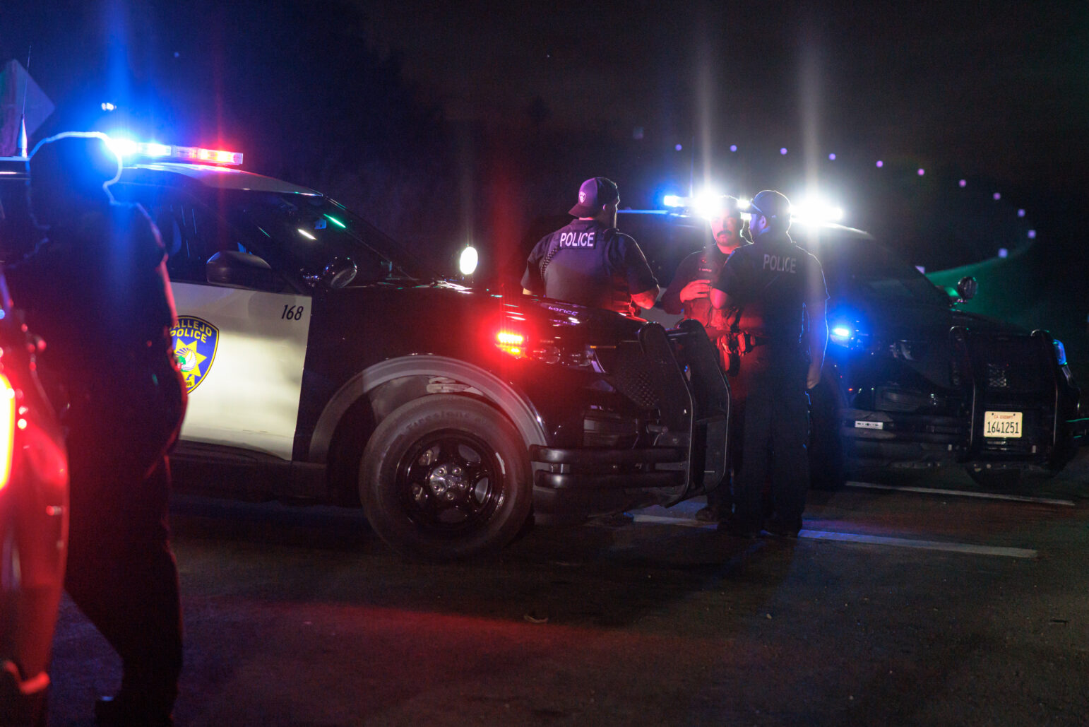 Multiple police vehicles with flashing lights surround a scene on a dark road. Several officers stand near the cars, talking amongst themselves, while the red and blue lights illuminate the night.