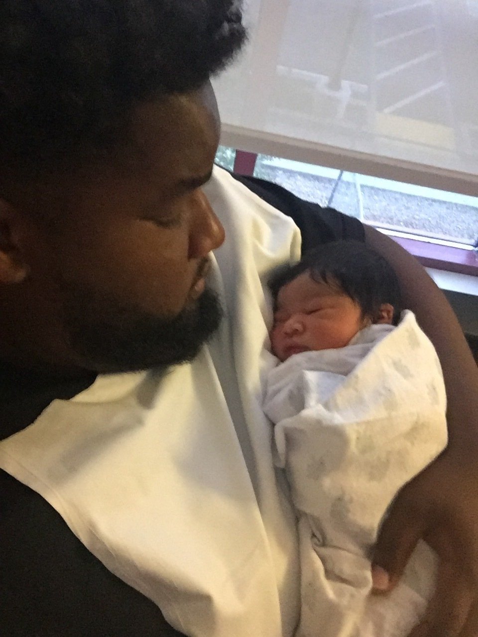 A tender moment between a father and his newborn child. The father, dressed in a black shirt, holds the sleeping baby swaddled in a light blanket against his chest. His gaze is affectionate and attentive as he watches over the infant, highlighting the deep connection and care in this familial bond.