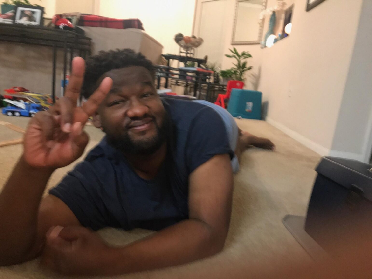 A cheerful man lying on the carpet inside a home gives a peace sign with his right hand. He wears a blue t-shirt and smiles warmly at the camera. The room around him is filled with children's toys and family photos, creating a casual and lived-in atmosphere.