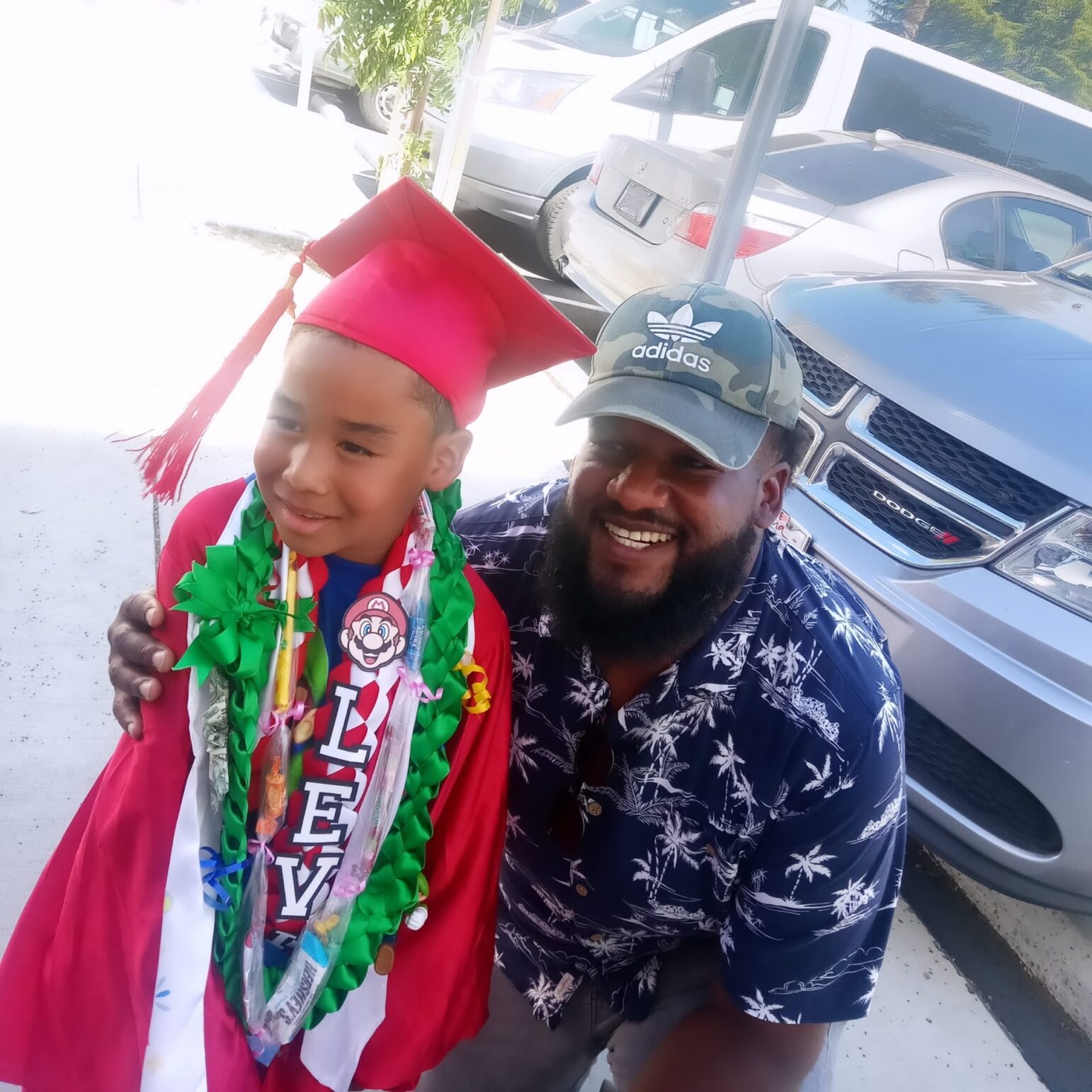 A young boy in a red graduation cap and gown, adorned with a lei and medals, smiles while standing next to a bearded man wearing a camouflage Adidas cap and a tropical print shirt. They are posing in a parking area with vehicles in the background.