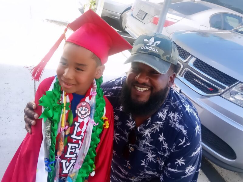 A young boy in a red graduation cap and gown, adorned with a lei and medals, smiles while standing next to a bearded man wearing a camouflage Adidas cap and a tropical print shirt. They are posing in a parking area with vehicles in the background.