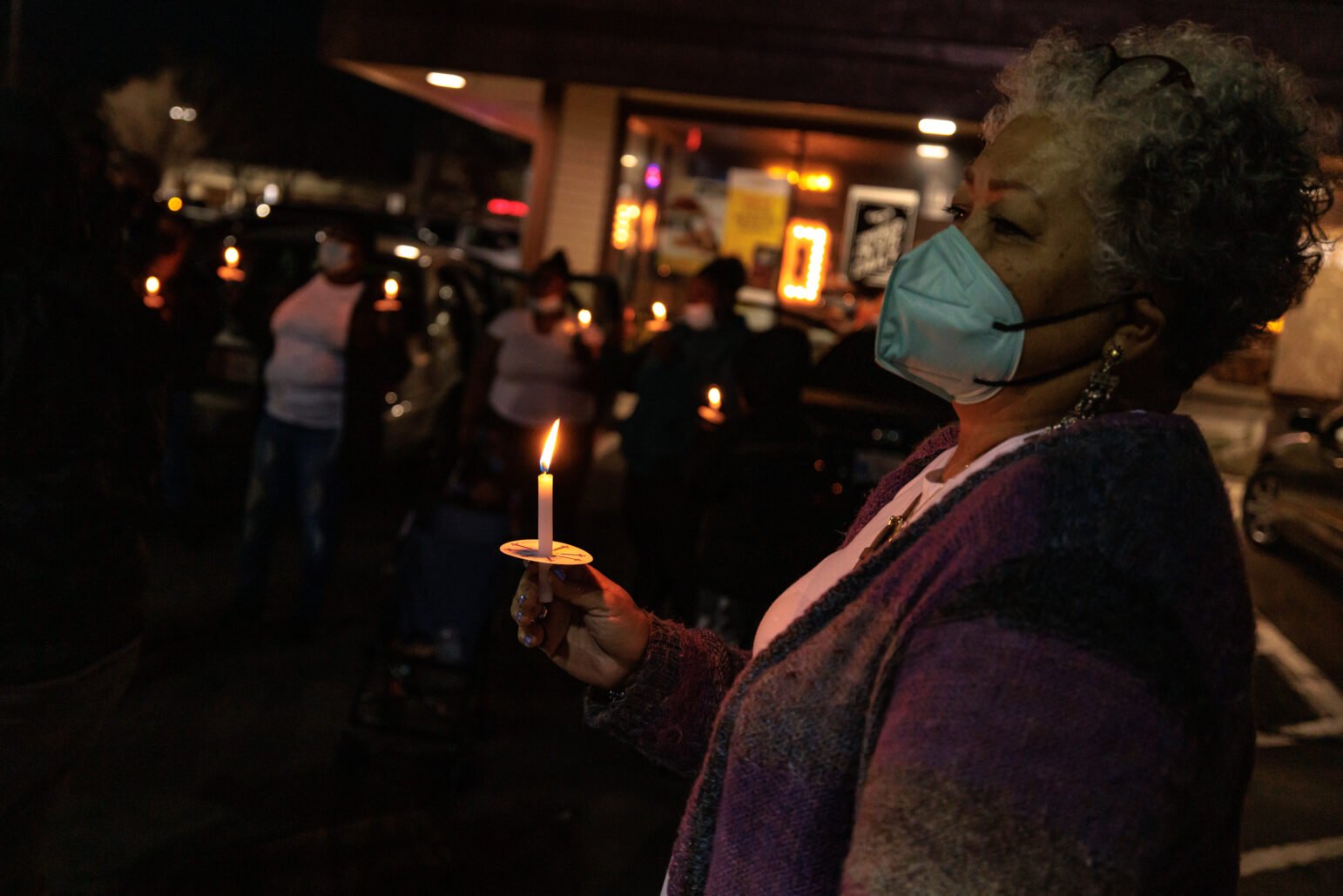 A woman in a mask holds a lit candle during an outdoor vigil at night. Other participants, also holding candles, are blurred in the background. The scene is solemn, with the soft glow of candlelight providing the primary source of illumination.