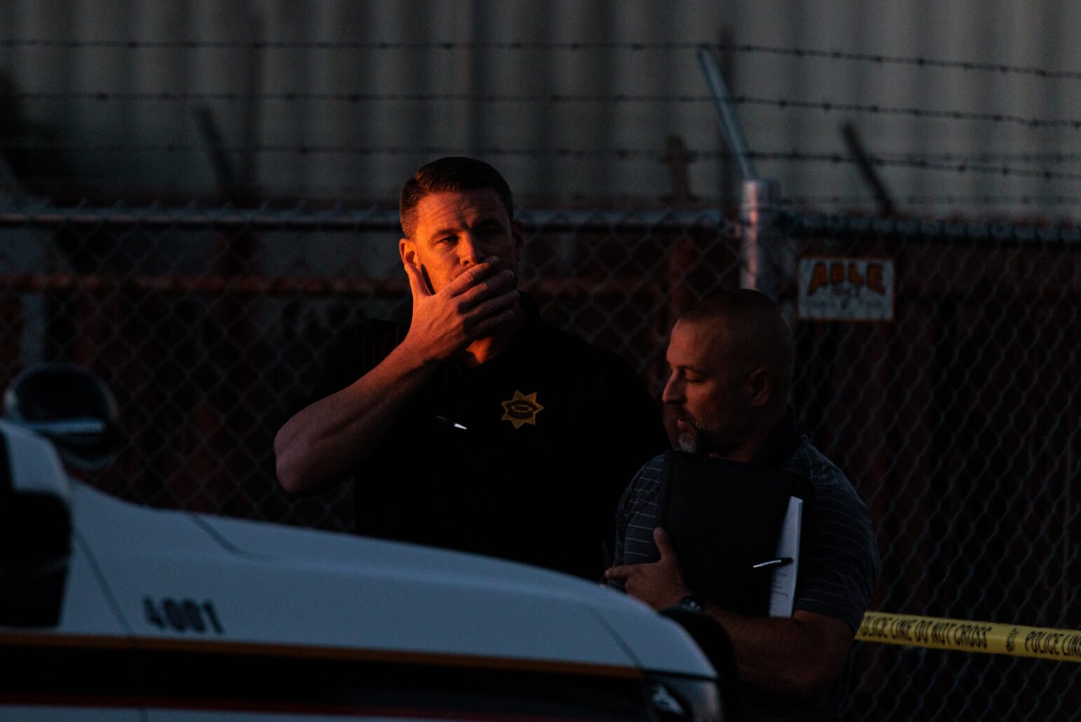 Two men, one in a Vallejo police polo and another in plain clothes, are captured during a somber moment. One of them holds his hand to his face in apparent distress or contemplation as they stand near a police vehicle at a crime scene, surrounded by caution tape and a chain-link fence.