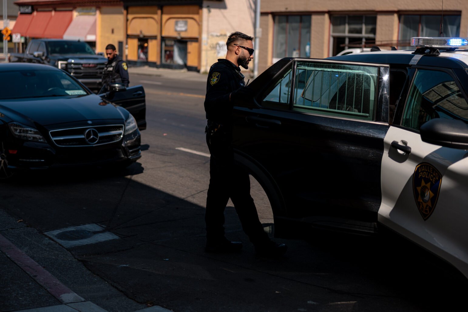 Two Vallejo police officers are seen during a traffic stop. One officer, standing near the back of a police vehicle, talks with a person in the backseat, while another stands nearby.