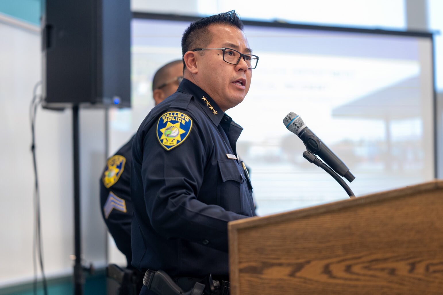 Vallejo Police Chief Jason Ta stands at a podium, speaking into a microphone. He is dressed in full police uniform, with his badge and department patch visible. Another officer stands behind him as they address a group at a public meeting.