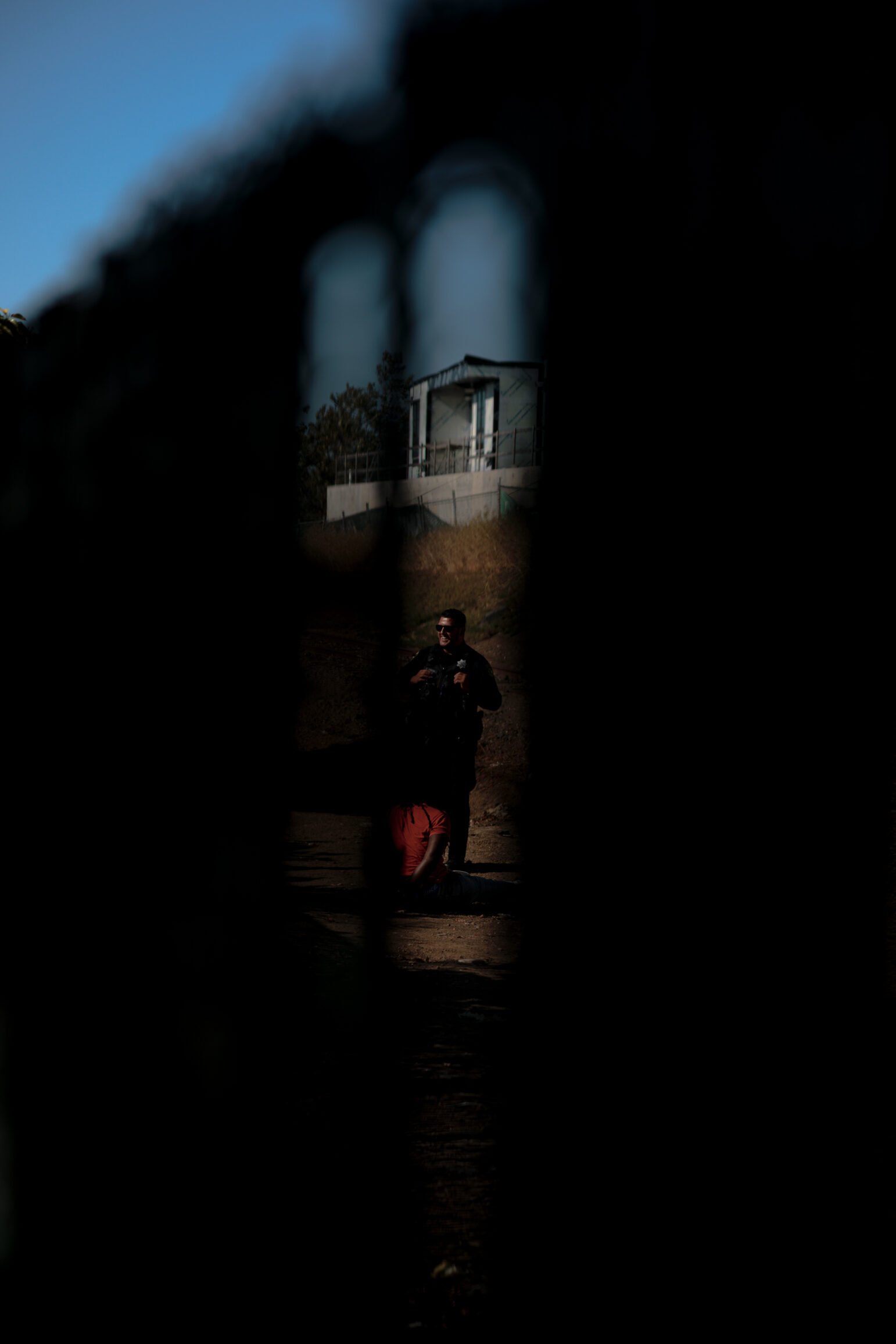 A shadowy figure of a smiling Vallejo police officer is visible through a narrow gap in a fence, observing a man seated on the ground in daylight. A structure is seen in the distance, adding to the sense of isolation and tension in the image.