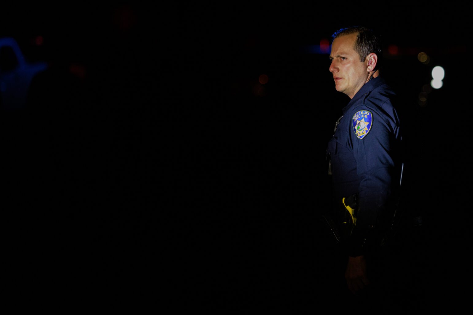 A Vallejo police officer in a dark uniform is partially illuminated in the night, standing with a stern expression. His badge and department patch are visible, though most of the image is shrouded in shadow.