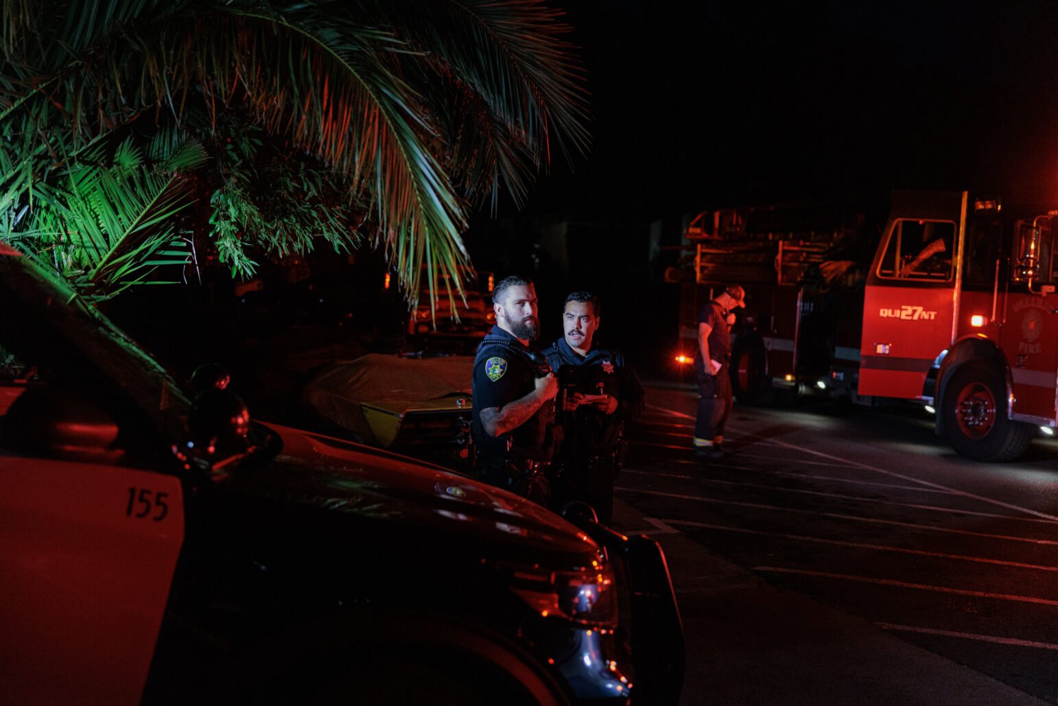 Two Vallejo police officers stand by a parked patrol vehicle under the glow of red emergency lights. A fire truck is visible in the background, with another person walking nearby. Palm fronds cast shadows on the officers and their surroundings.