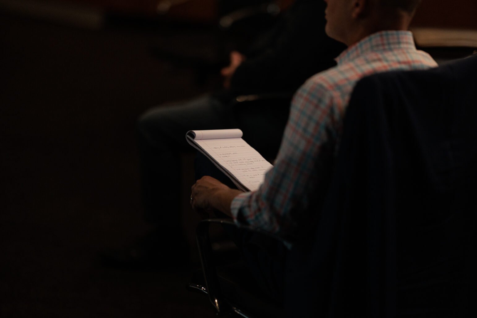 A man in a plaid shirt takes notes in a city council chambers, focusing on his notepad during a public meeting. The dark background obscures other seated individuals, drawing attention to the notepad.