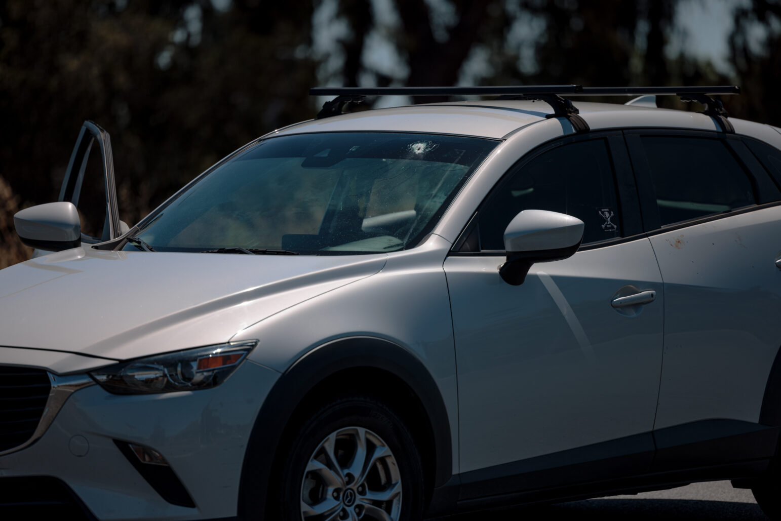 A silver SUV with its driver's side door slightly ajar is shown in the image. A bullet hole is visible in the windshield, indicating damage from a shooting. There are faint marks, possibly blood, near the door handle on the right side of the vehicle. The car is parked in an outdoor setting under sunlight, with trees blurred in the background.