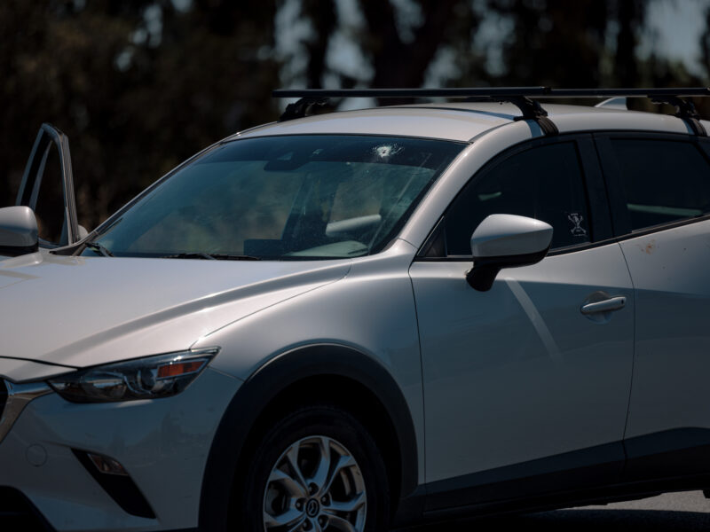 A silver SUV with its driver's side door slightly ajar is shown in the image. A bullet hole is visible in the windshield, indicating damage from a shooting. There are faint marks, possibly blood, near the door handle on the right side of the vehicle. The car is parked in an outdoor setting under sunlight, with trees blurred in the background.