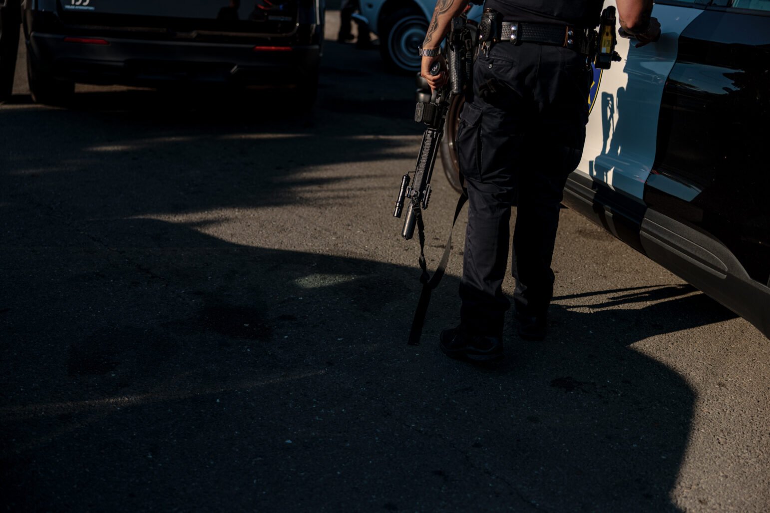 A Vallejo police officer, seen from the waist down, is holding a rifle while standing next to a patrol car. The shadow of his figure and the vehicle dominate the lower portion of the frame.