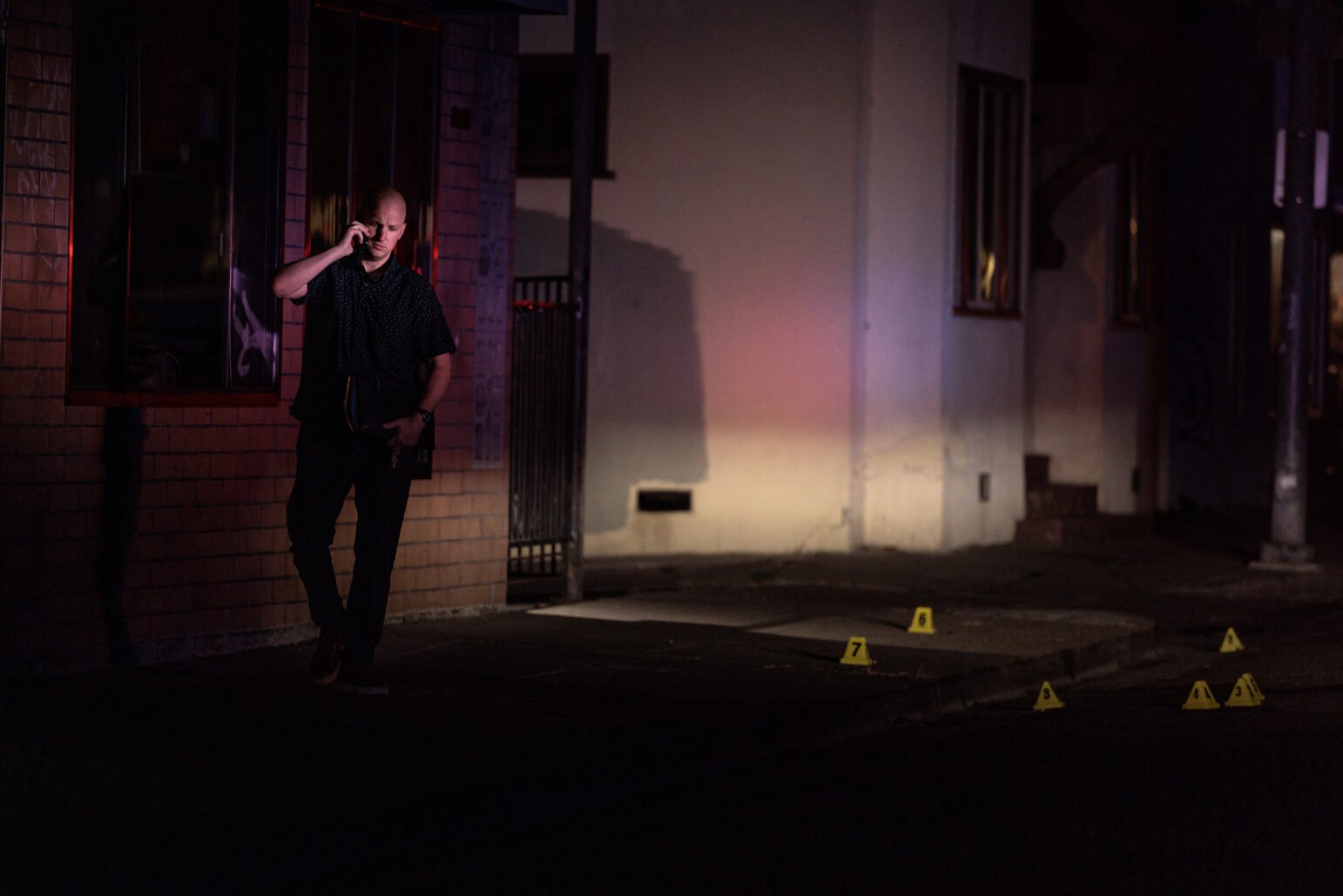 A police detective stands outside a building at night, illuminated by the glow of streetlights and emergency vehicle lights. He holds a phone to his ear while placing his hand on his hip. Yellow evidence markers labeled 3, 4, 6, 7, and 9 are scattered on the sidewalk and street, denoting a crime scene investigation. The scene is quiet, with shadows cast on the building's brick wall and windows, adding to the somber atmosphere.