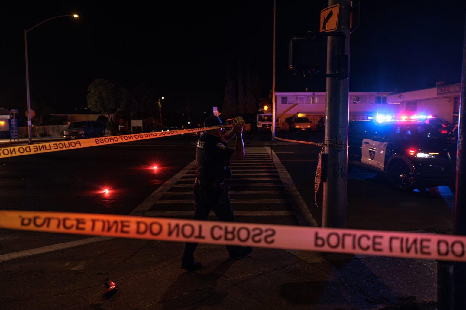 A Vallejo police officer secures a scene by taping up "Police Line Do Not Cross" barriers at an intersection during the night. Red flares on the street add urgency to the scene, while police vehicles with flashing lights sit in the background.