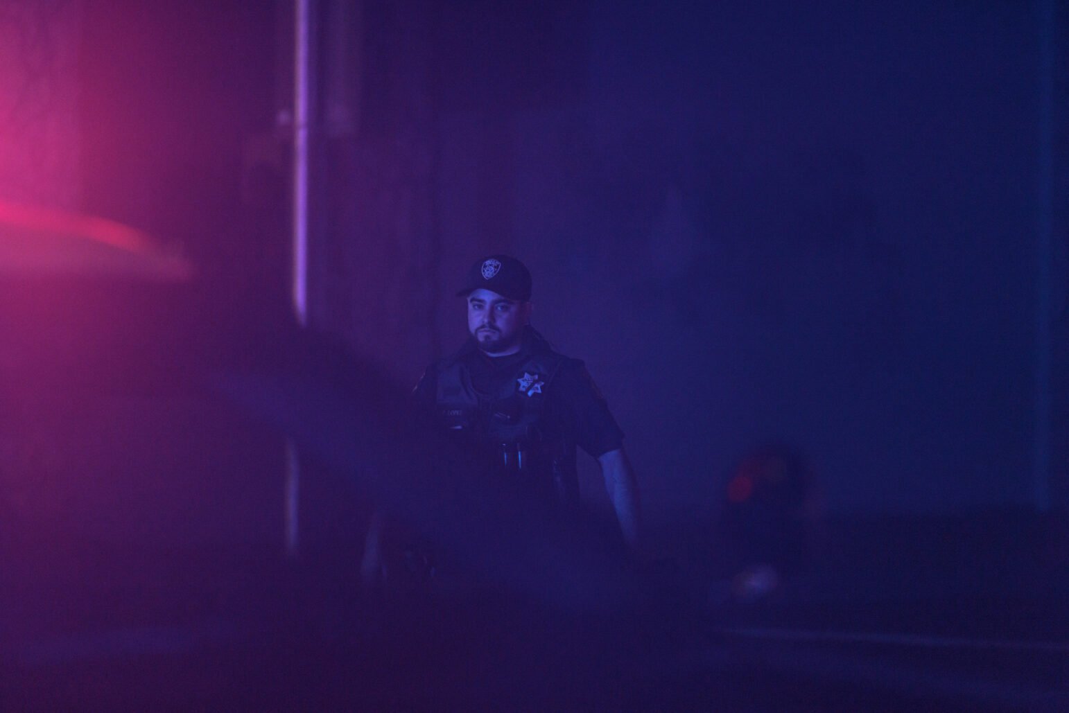 A Vallejo police officer is bathed in red and blue light during nighttime. His focused expression is captured as he observes the scene ahead of him, with the light reflecting off his uniform and surroundings.