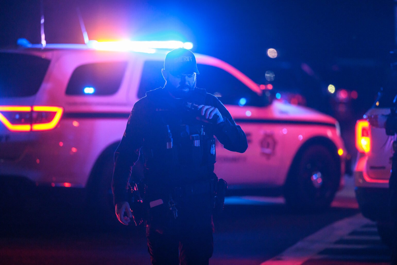 A Solano County Sheriff's Office deputy walks toward the camera in low light, illuminated by the flashing red and blue lights of a police vehicle parked behind him. The deputy wears a tactical vest and holds a radio to his mouth, appearing to communicate. The background is dark, with the bright lights of police vehicles casting colorful reflections across the scene, highlighting the tense and active nature of the situation.