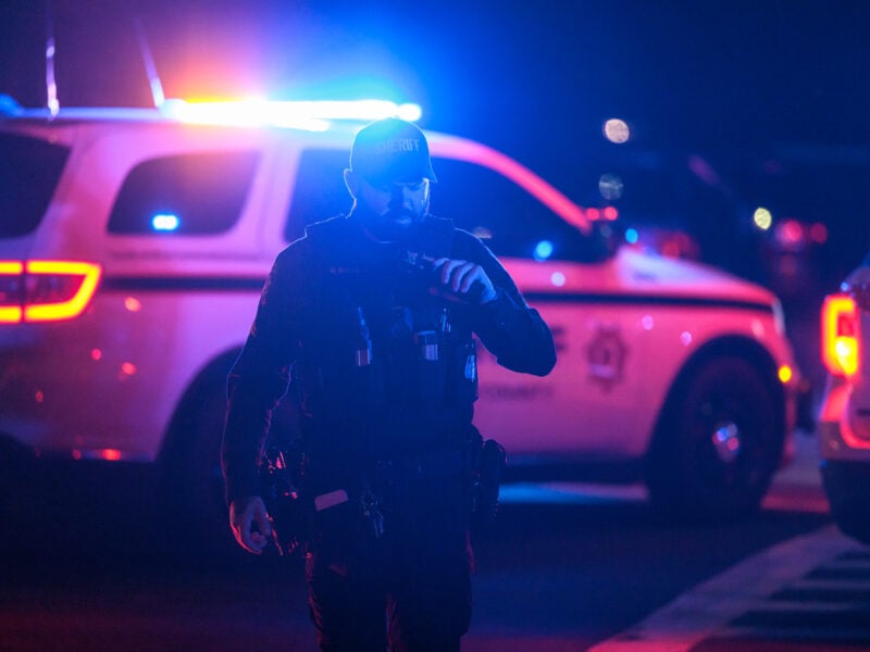 A Solano County Sheriff's Office deputy walks toward the camera in low light, illuminated by the flashing red and blue lights of a police vehicle parked behind him. The deputy wears a tactical vest and holds a radio to his mouth, appearing to communicate. The background is dark, with the bright lights of police vehicles casting colorful reflections across the scene, highlighting the tense and active nature of the situation.