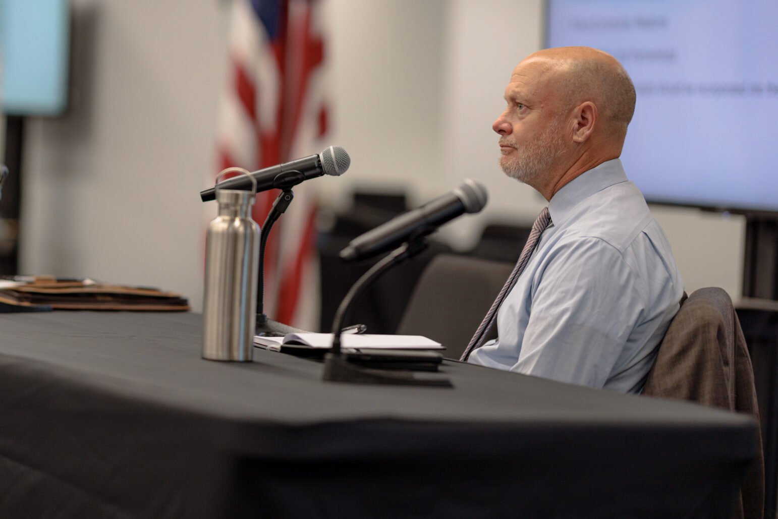 Vallejo City Manager Andrew Murray sits at a table during a public meeting, with a microphone in front of him. The background shows an American flag, and the atmosphere is formal, suggesting an official setting.