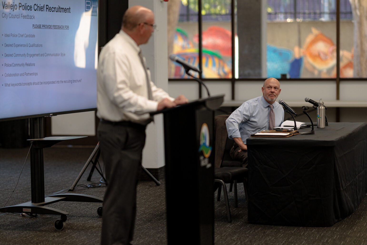 A speaker stands at a podium, addressing a meeting about Vallejo Police Chief recruitment. A screen to the left displays discussion points, while another man seated at a table listens attentively, with microphones and papers in front of him. The window in the background shows a colorful mural, brightening the formal setting.