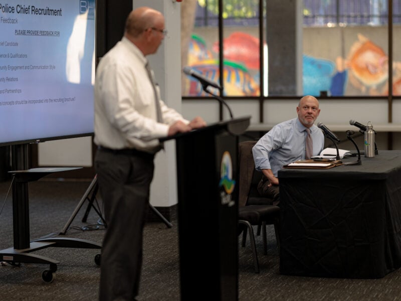 A speaker stands at a podium, addressing a meeting about Vallejo Police Chief recruitment. A screen to the left displays discussion points, while another man seated at a table listens attentively, with microphones and papers in front of him. The window in the background shows a colorful mural, brightening the formal setting.