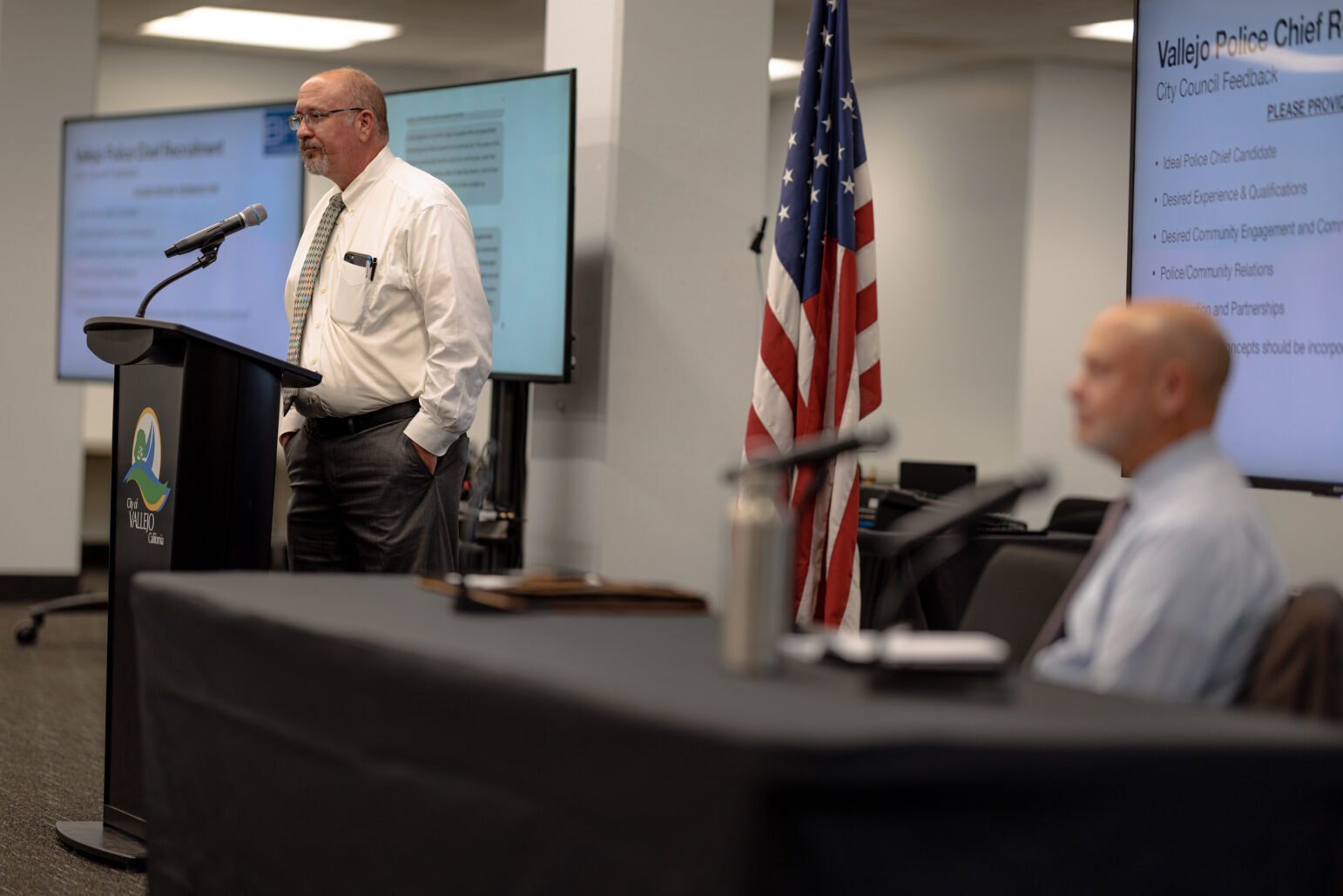 A man in a white shirt and tie stands at a podium with the City of Vallejo logo, delivering a presentation. Behind him, an American flag is displayed, and a large screen shows feedback topics related to the recruitment of a new police chief, such as qualifications and community relations. A seated man in a blue shirt, slightly out of focus, is positioned in the foreground, listening to the speaker. The setting appears to be a community meeting room.