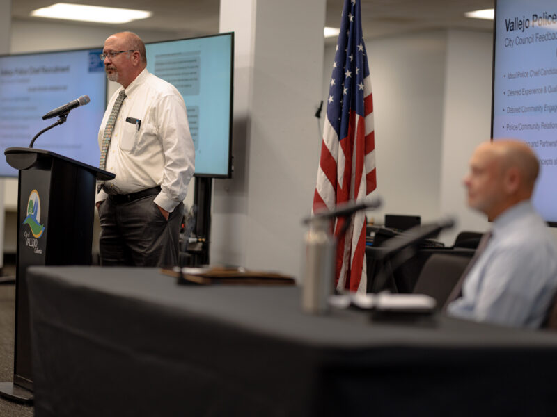 A man in a white shirt and tie stands at a podium with the City of Vallejo logo, delivering a presentation. Behind him, an American flag is displayed, and a large screen shows feedback topics related to the recruitment of a new police chief, such as qualifications and community relations. A seated man in a blue shirt, slightly out of focus, is positioned in the foreground, listening to the speaker. The setting appears to be a community meeting room.