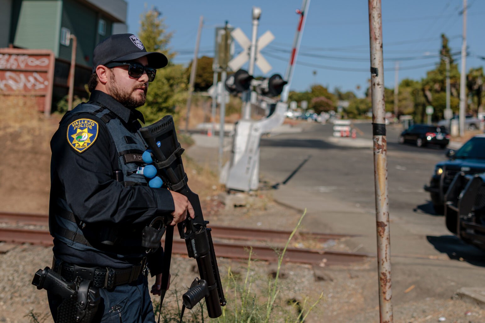 A Vallejo police officer wearing sunglasses and tactical gear stands near a railroad crossing, holding a less-lethal launcher. The background includes the crossing gate, urban surroundings, and a distant street with cars.