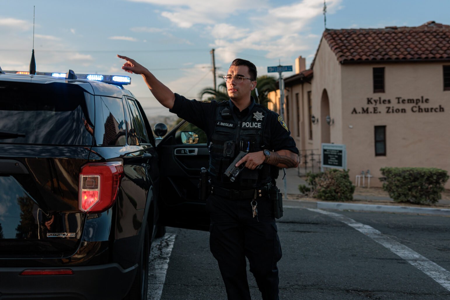 A Vallejo police officer gestures while holding a gun in one hand, his uniform and vehicle illuminated by the warm light of late afternoon. The background shows a sign for Kyles Temple A.M.E. Zion Church.