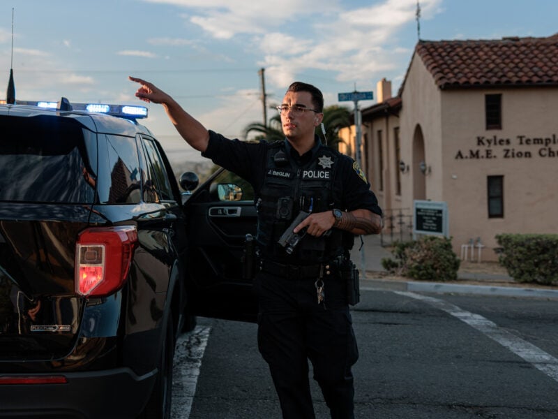 A Vallejo police officer gestures while holding a gun in one hand, his uniform and vehicle illuminated by the warm light of late afternoon. The background shows a sign for Kyles Temple A.M.E. Zion Church.