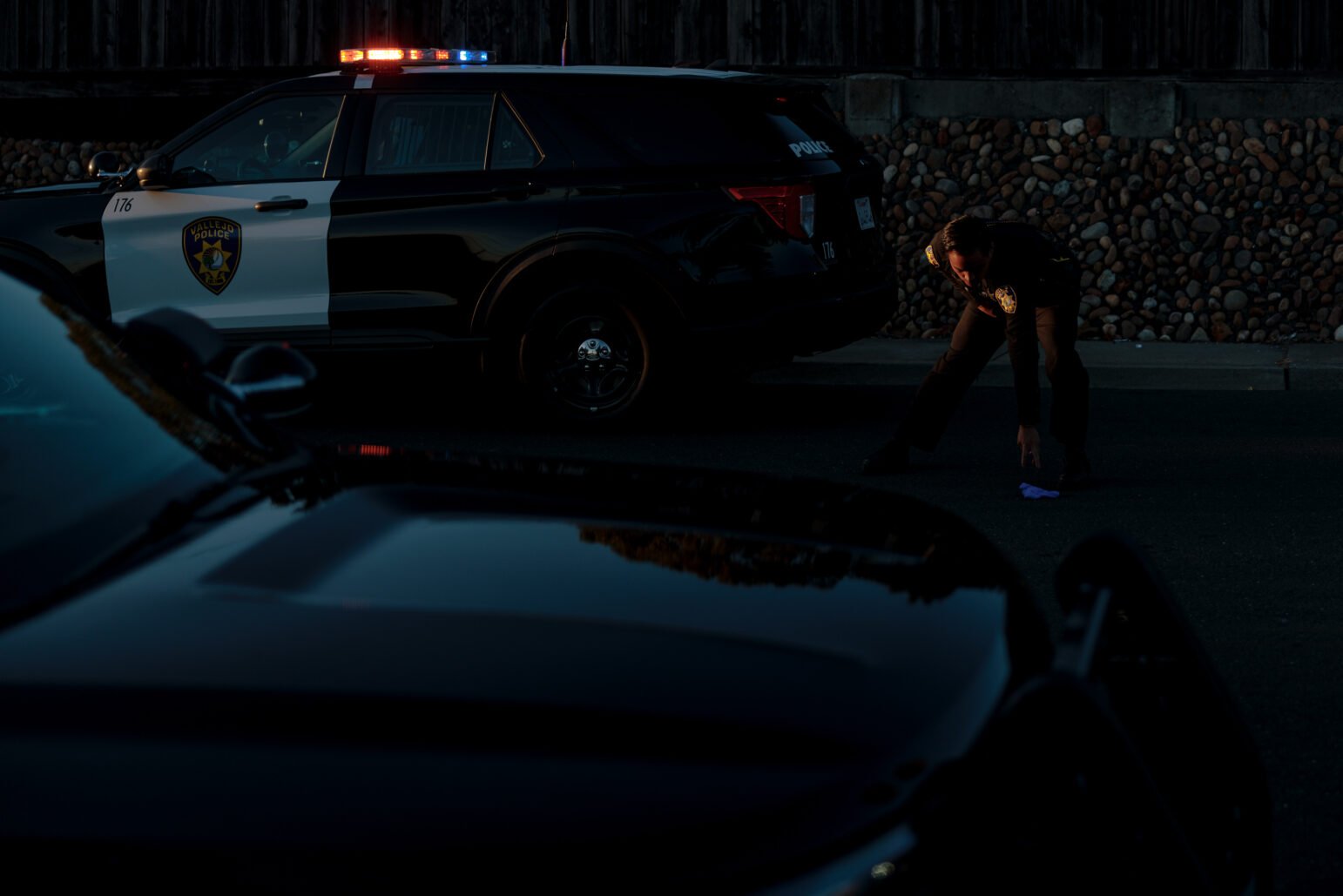 A Vallejo police officer crouches down in front of a black police SUV to pick up an item from the ground, which appears to be a piece of evidence. Police lights reflect off the dark surfaces of the vehicles around the officer.