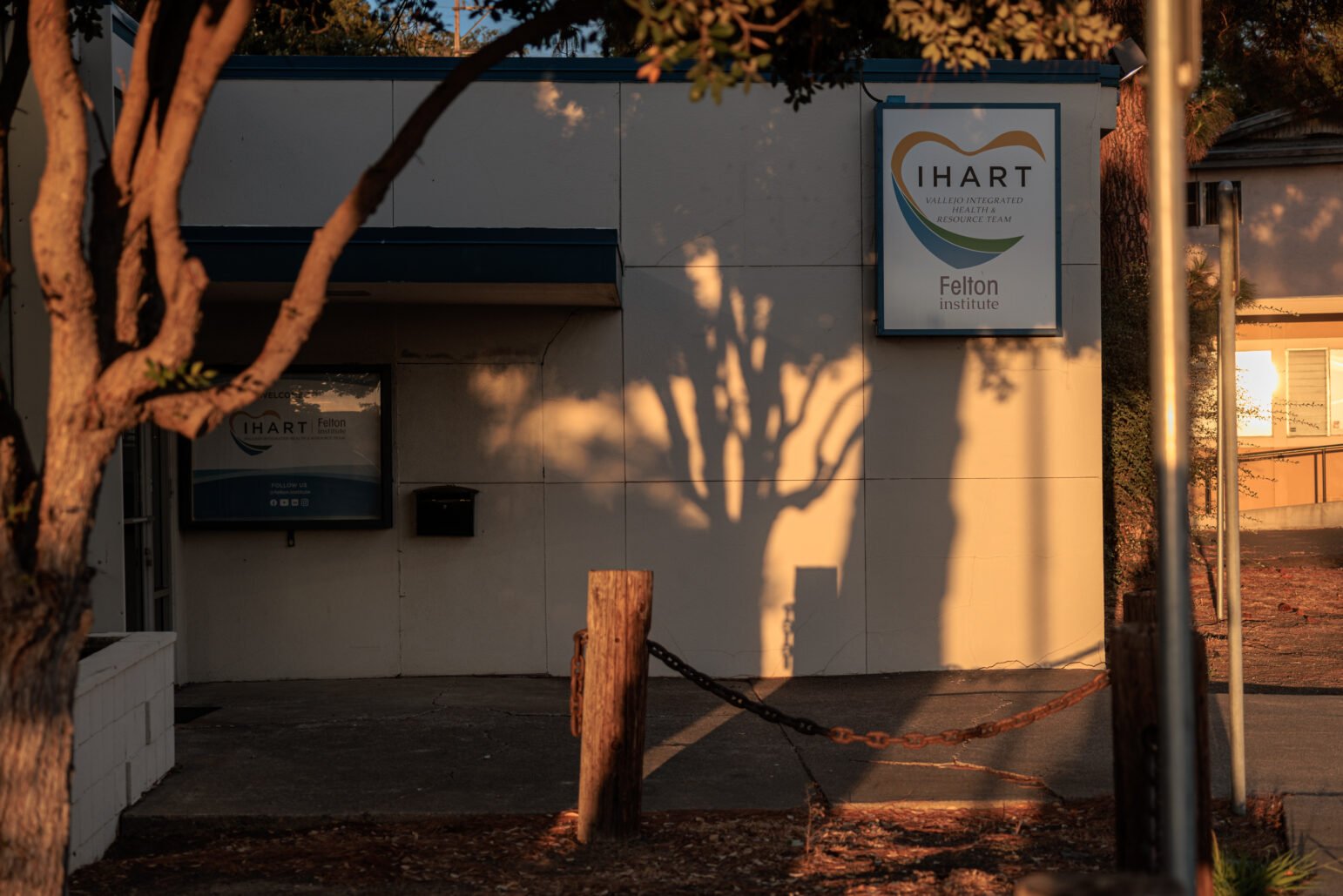 The exterior of the IHART (Vallejo Integrated Health Resource Team) building is shown during sunset. Shadows of trees stretch across the front of the building, with the Felton Institute sign prominently displayed near the entrance, casting a warm, calm atmosphere.