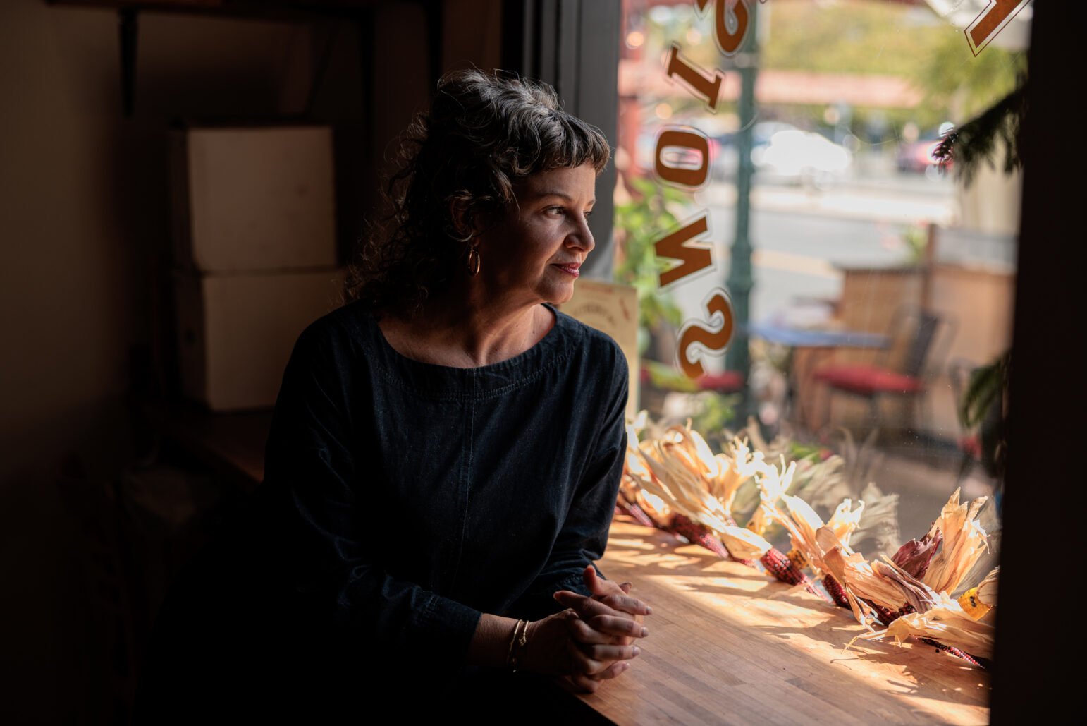 A woman with curly hair sits inside a cafe, gazing thoughtfully out of a large window. The soft sunlight highlights her face as she rests her hands on a table. The cafe window displays decorative elements, and a quiet street scene is visible outside.