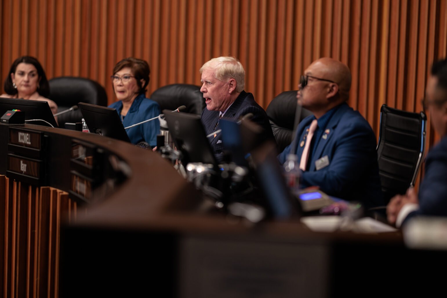 Vallejo Mayor Robert McConnell speaks passionately into a microphone during a public meeting, seated among other council members at a curved desk. Two women and two men, including a man in glasses and a blue suit, are focused on the proceedings. The setting appears formal, with the panel framed by wood paneling and computer monitors.