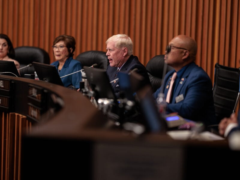 Vallejo Mayor Robert McConnell speaks passionately into a microphone during a public meeting, seated among other council members at a curved desk. Two women and two men, including a man in glasses and a blue suit, are focused on the proceedings. The setting appears formal, with the panel framed by wood paneling and computer monitors.