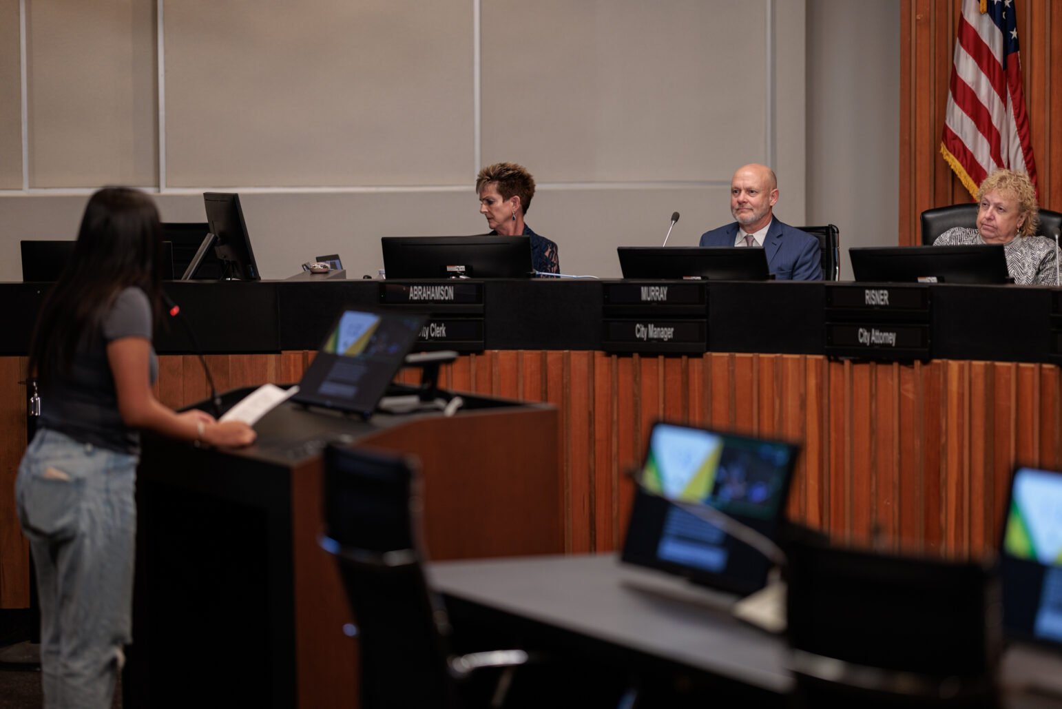 A young woman stands at a podium, addressing Vallejo's city council, city manager, city clerk and city attorney. The setting is a formal council chamber, with nameplates and an American flag visible, emphasizing the official nature of the meeting.