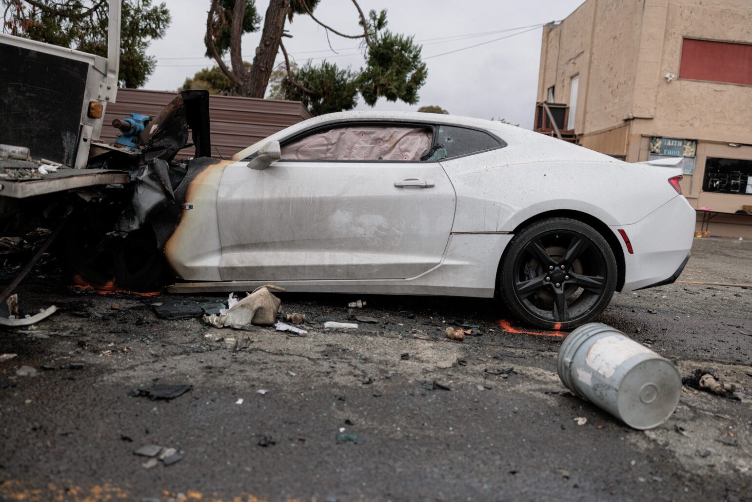 A white Chevrolet Camaro with severe front-end damage is seen after a collision with a utility truck. The car’s windshield is shattered, and airbags have deployed. A police officer inspects the wreckage, with debris scattered around the scene.